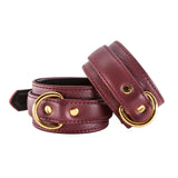 The JT Signature Collection Wrist Restraints are shown against a blank background. The cuffs are made of Bordeaux leather with a black interior lining. The cuffs have a D-ring and a buckle closure. The hardware is gold.