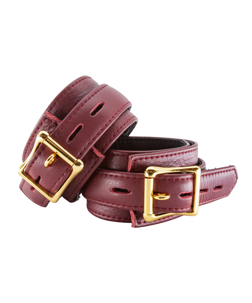 The JT Signature Collection Wrist Restraints are shown from the back against a blank background. The cuffs are made of Bordeaux leather with a black interior lining. The cuffs have a D-ring and a buckle closure. The hardware is gold.