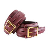 The JT Signature Collection Wrist Restraints are shown from the back against a blank background. The cuffs are made of Bordeaux leather with a black interior lining. The cuffs have a D-ring and a buckle closure. The hardware is gold.