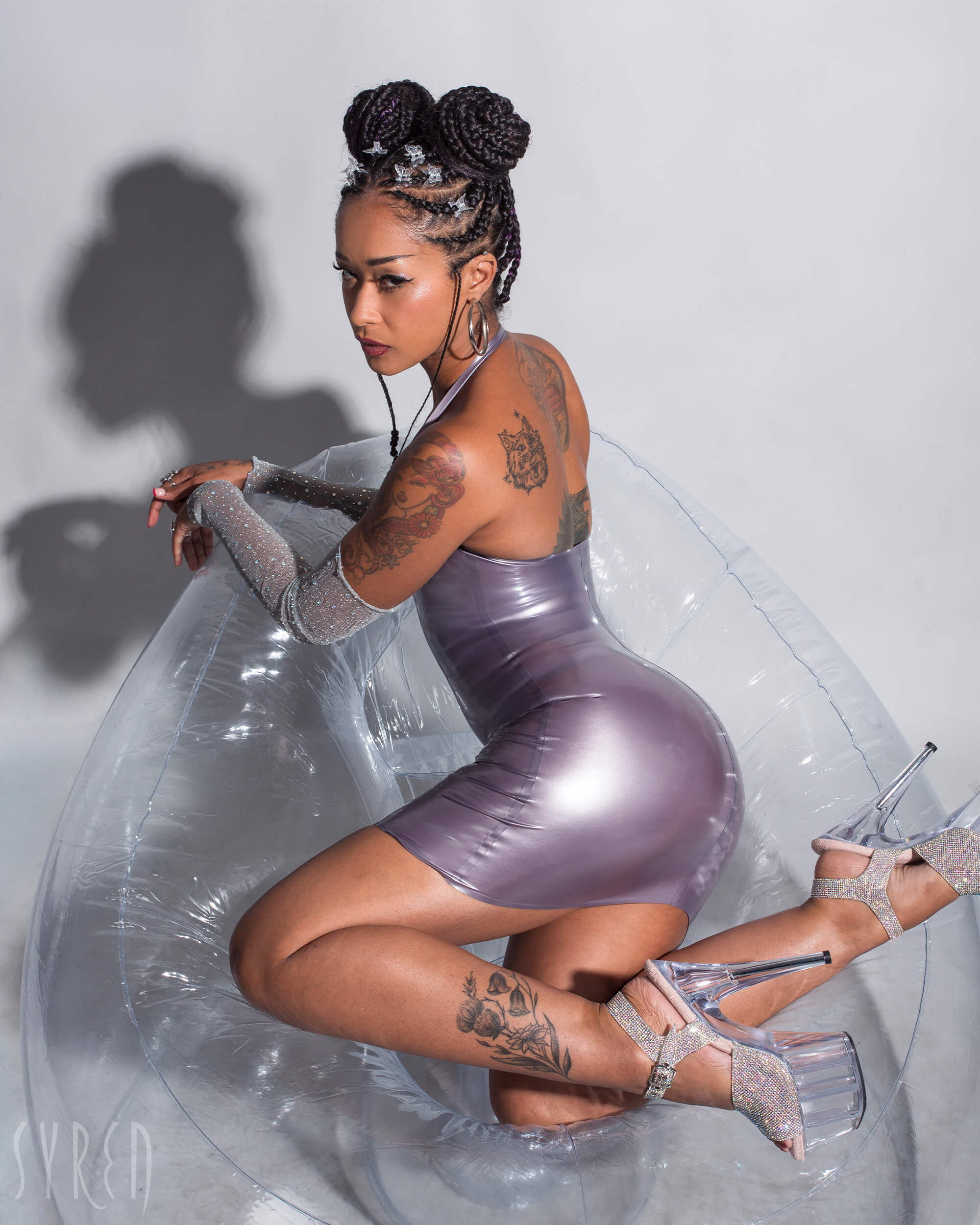 A model is shown sitting on a clear inflatable chair wearing the Syren Latex pearl Lilac halter dress against a light gray background.