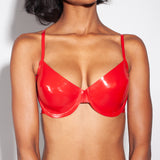 A close-up of a model is shown wearing the Syren latex Mila bra in red. She is standing in front of a light gray background.