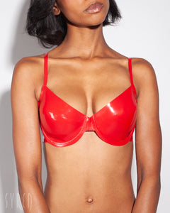 A close-up of a model is shown wearing the Syren latex Mila bra in red. She is standing in front of a light gray background.