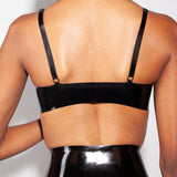 A close-up of a model is shown from the back wearing the Syren latex Mila bra in black. She is standing in front of a light gray background