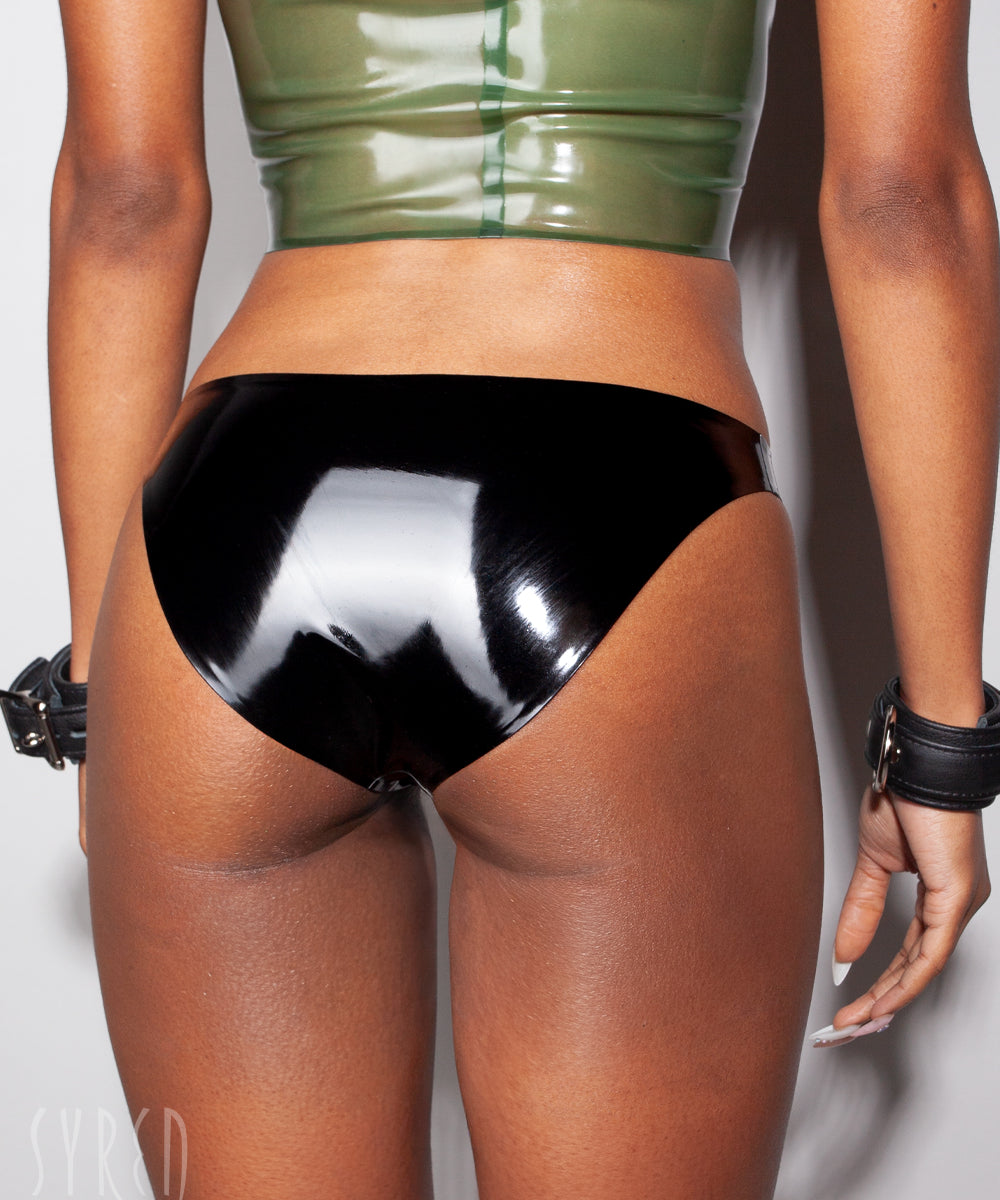 A close-up of a models waist is shown wearing the Syren latex mila panty in black. She is standing in front of a light gray background. 