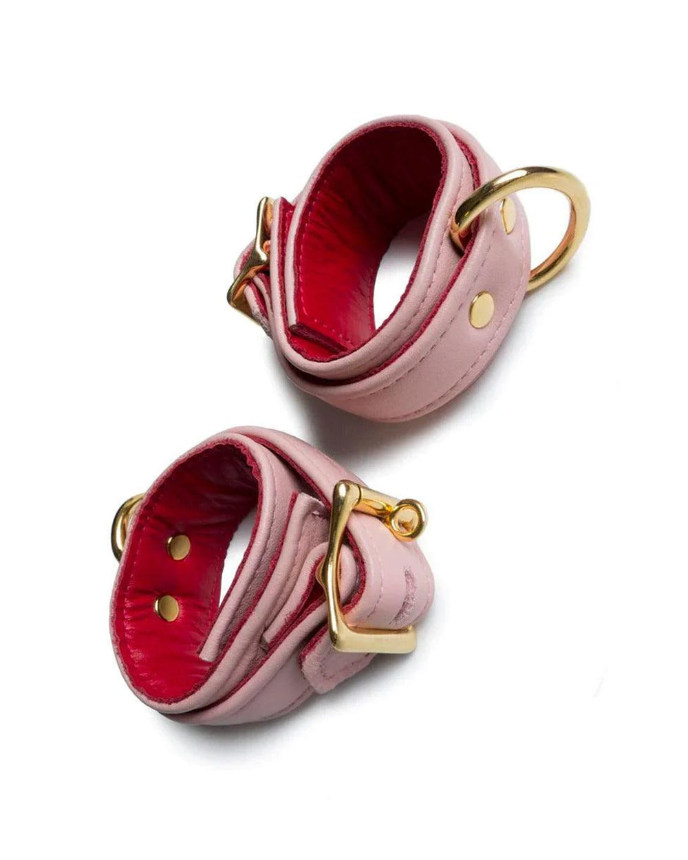 A pair of Premium Garment Leather Wrist Cuffs With 18 Karat Gold Plated Hardware is shown against a blank background. The cuffs are light pink with red interior lining. The buckles, D-rings, and rivets are all gold.