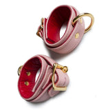 A pair of Premium Garment Leather Wrist Cuffs With 18 Karat Gold Plated Hardware is shown against a blank background. The cuffs are light pink with red interior lining. The buckles, D-rings, and rivets are all gold.
