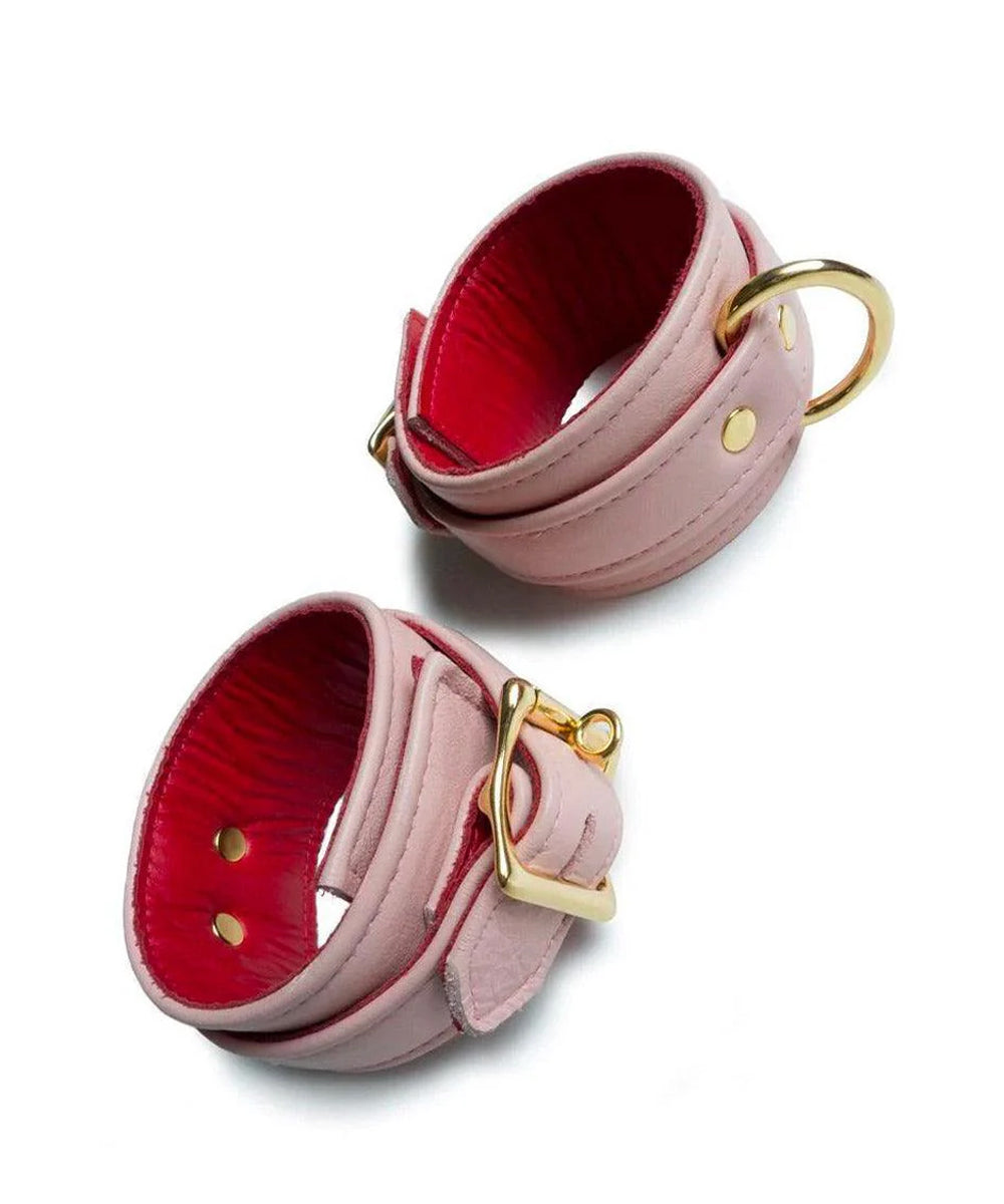 A set of Premium Garment Leather Ankle Cuffs With 18k Gold Plated Hardware are displayed against a blank leather background. The cuffs are made of light pink leather with a red inner lining. They each have one D-ring and fasten with a lockable buckle.