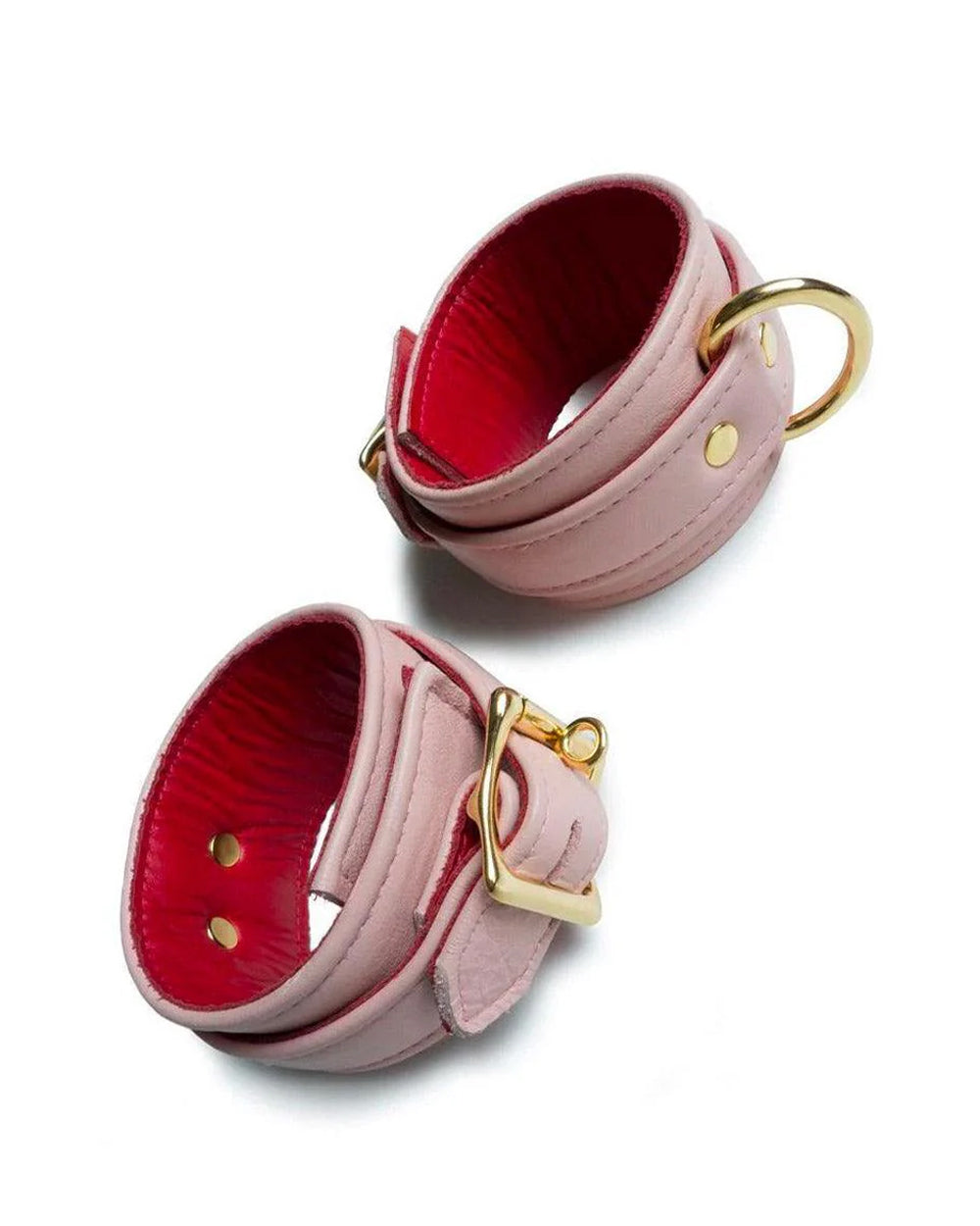 A set of Premium Garment Leather Ankle Cuffs With 18k Gold Plated Hardware are displayed against a blank leather background. The cuffs are made of light pink leather with a red inner lining. They each have one D-ring and fasten with a lockable buckle.