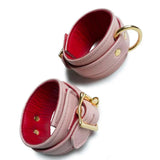 A set of Premium Garment Leather Ankle Cuffs With 18k Gold Plated Hardware are displayed against a blank leather background. The cuffs are made of light pink leather with a red inner lining. They each have one D-ring and fasten with a lockable buckle.