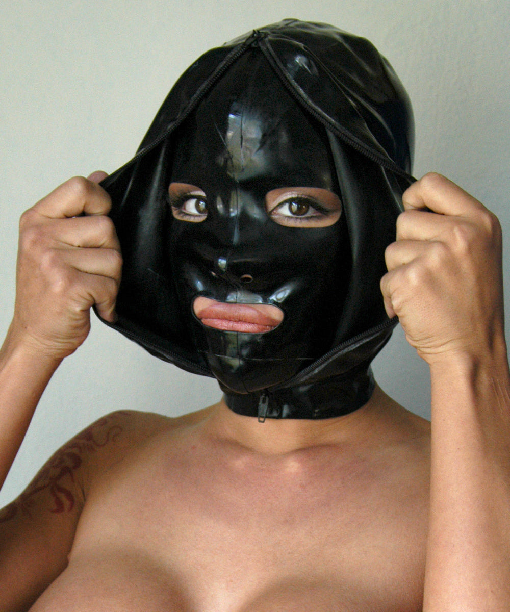 A close-up of a woman's face is shown wearing the black Extreme Rubber Hood. The hood is unzipped, and she pulls apart the outer layers to reveal the bank robber-style mask underneath.