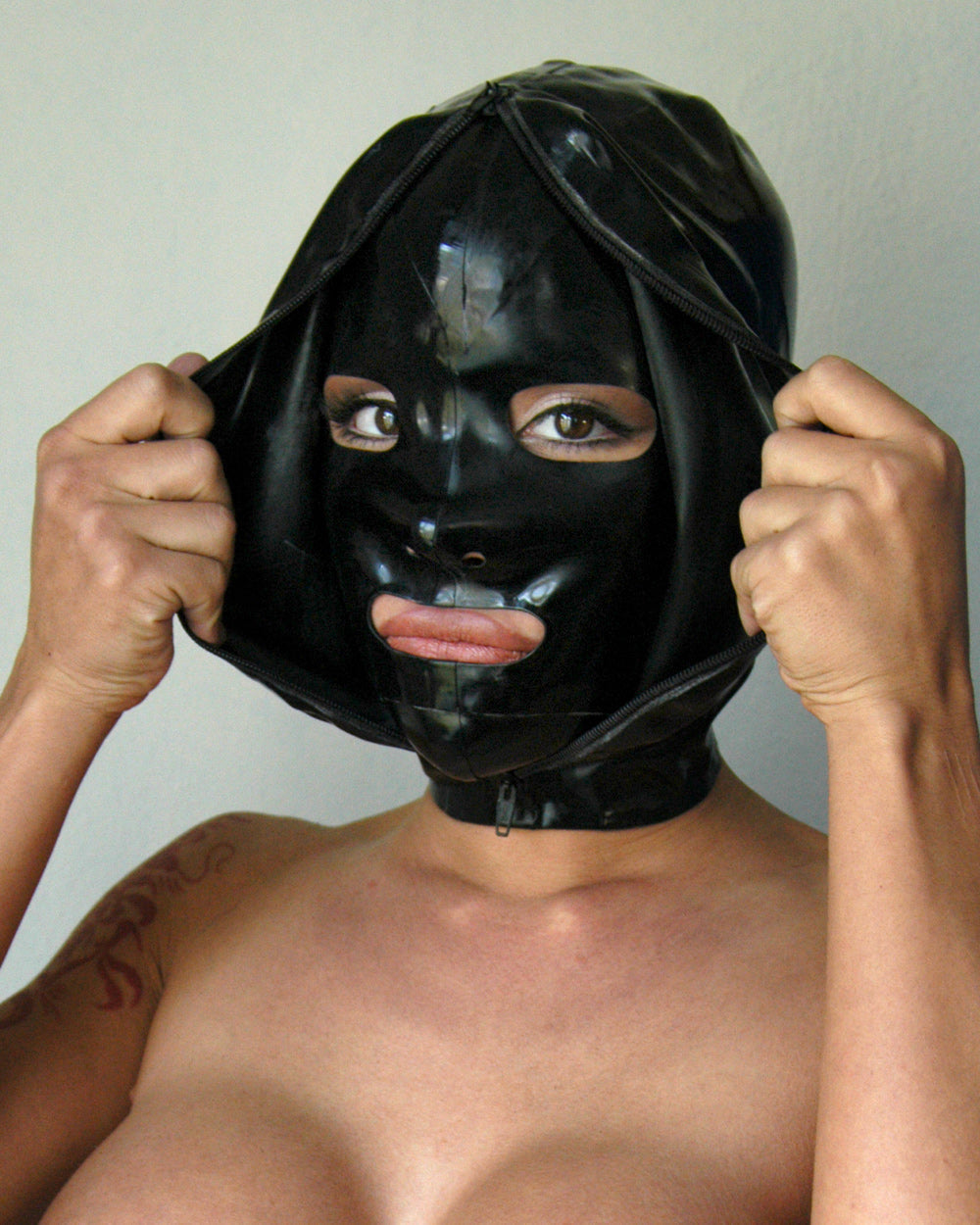A close-up of a woman's face is shown wearing the black Extreme Rubber Hood. The hood is unzipped, and she pulls apart the outer layers to reveal the bank robber-style mask underneath.