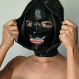 A close-up of a woman's face is shown wearing the black Extreme Rubber Hood. The hood is unzipped, and she pulls apart the outer layers to reveal the bank robber-style mask underneath.